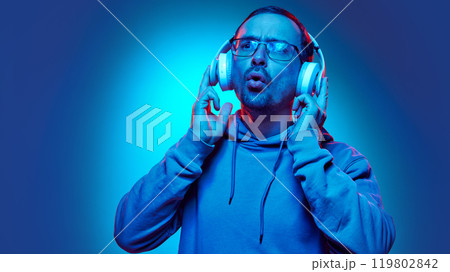 Young man listening music and sig with joy in neon light against gradient studio background. Hobby. 119802842