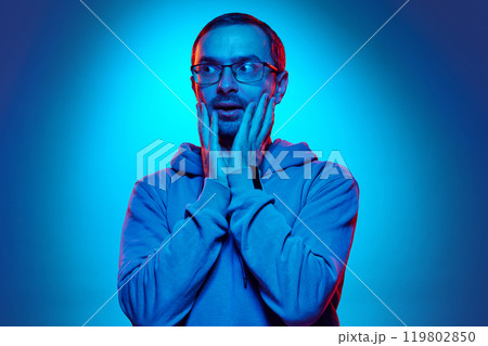 Portrait of young man dressed casual hoodie and holding hands near mouth as is he scared in neon light against gradient studio background. 119802850