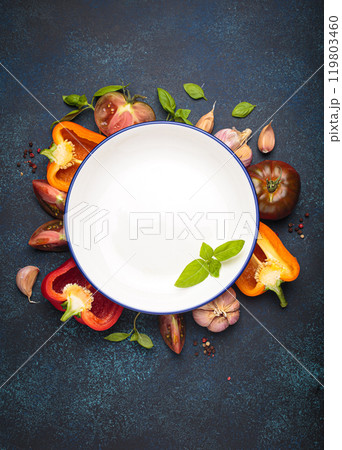 Empty white plate surrounded by fresh cooking ingredients and vegetables on dark blue background, space for text 119803460
