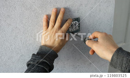 a worker repairs an electric socket or light switch by screwing a screw into it with a screwdriver against a background of gray wallpaper 119803525