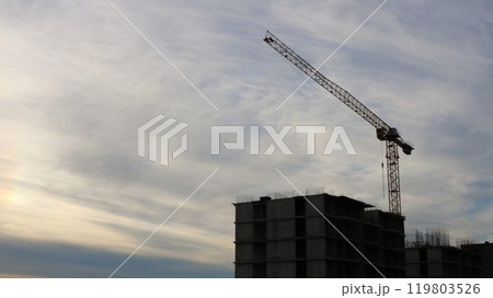 Behind an unfinished multi-story building stands a construction crane against a yellow-blue cloudy sky 119803526