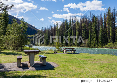 A picnic table and benches on the green meadow next to a river. Mountain and forest in the background. Maligne River in Jasper National Park, Alberta, Canada. Canadian Rockies. A picnic table and benches on the green meadow next to a river. Mountain and forest in the background. Maligne River in Jasper National Park, Alberta, Canada. Canadian Rockies. 119803871