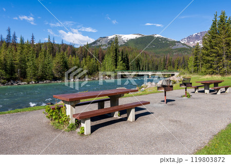 A picnic table and benches on the green meadow next to a river. Mountain and forest in the background. Maligne River in Jasper National Park, Alberta, Canada. Canadian Rockies. A picnic table and benches on the green meadow next to a river. Mountain and forest in the background. Maligne River in Jasper National Park, Alberta, Canada. Canadian Rockies. 119803872