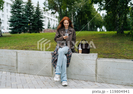 Portrait of happy young woman sitting on a bench enjoying beautiful day at park, using smartphone and drinking coffee during break. Stylish redhead woman relax alone, enjoying free time in outdoors. 119804941