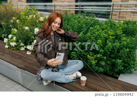 Cheerful redhead female taking selfie with smartphone, experimenting with various poses and expressions in urban setting filled with greenery and modern architecture, enjoying free time in city. 119804942