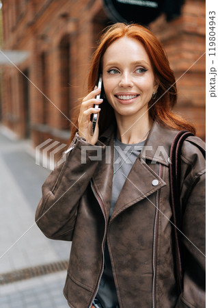 Vertical shot of cheerful young woman strolling down city street, enjoying pleasant conversation on smartphone. Happy redhead female in stylish leather jacket reflect carefree urban lifestyle. 119804943
