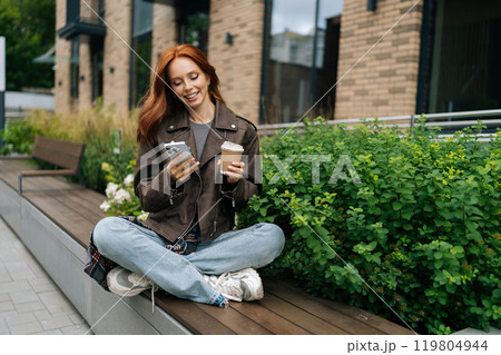 Portrait of joyful redhead female in jacket sitting on urban bench, enjoying cup of coffee and using mobile phone, taking break from day to relax and catch up with friends on summer day outdoors. 119804944