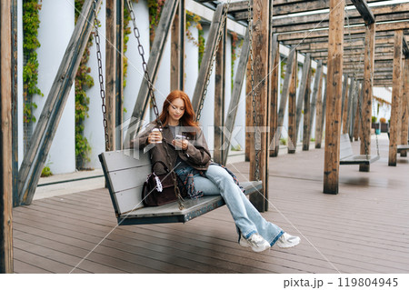Wide shot of relaxed female with smile on face swinging on swings in city park, drinking coffee and browsing smartphone, exuding tranquility. Happy woman enjoying coffee on swing, using typing phone. 119804945