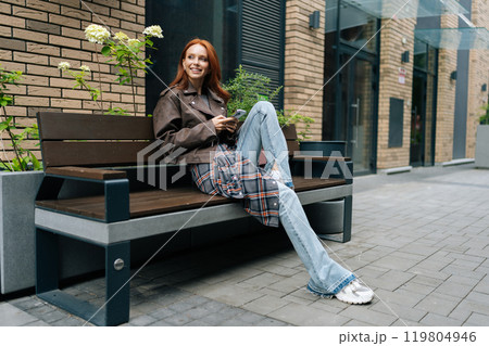 Pretty redhead female browsing smartphone, enjoying beautiful day in city. Happy red-haired young woman in leather jacket sitting on bench in city, holding using mobile phone outdoors. 119804946