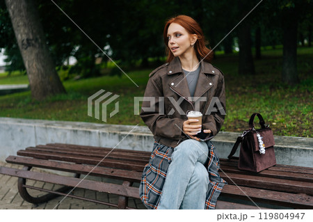 Portrait of serene young woman with long red hair enjoying peaceful moment outdoors, sipping coffee sitting on bench in city park, surrounded by greenery, looking stylish on autumn day. 119804947