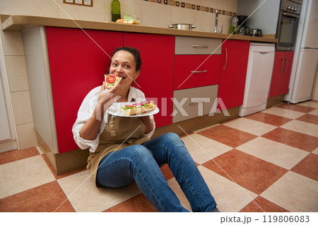Woman enjoying homemade sandwiches sitting on kitchen floor Woman enjoying homemade sandwiches sitting on kitchen floor 119806083