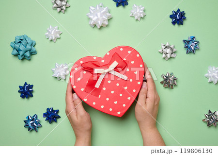 Female hands holding heart shaped gift box and bows on green background, top view Female hands holding heart shaped gift box and bows on green background, top view 119806130
