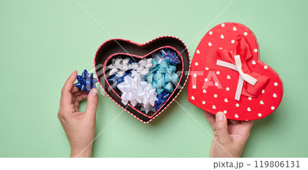 Female hands holding heart shaped gift box and bows on green background, top view Female hands holding heart shaped gift box and bows on green background, top view 119806131
