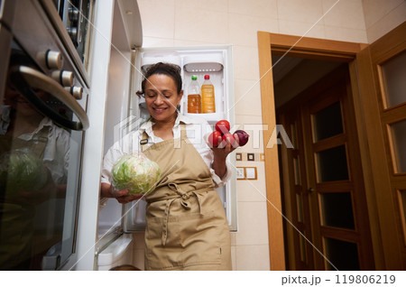 Woman in kitchen holding fresh vegetables by open fridge 119806219