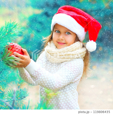 Christmas smiling child little girl in santa red hat holding ball near a branch tree 119806435