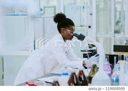 Scientist wearing safety gear and white lab coat observing through microscope while conducting research in laboratory surrounded by various scientific equipment and chemical solutions Scientist wearing safety gear and white lab coat observing through microscope while conducting research in laboratory surrounded by various scientific equipment and chemical solutions 119808000