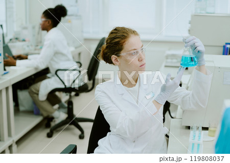 Scientist holding blue chemical solution in laboratory while a colleague working in background, both focused on tasks and using advanced equipment surrounded by scientific tools Scientist holding blue chemical solution in laboratory while a colleague working in background, both focused on tasks and using advanced equipment surrounded by scientific tools 119808037