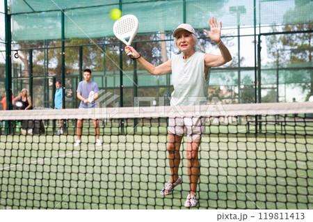 Concentrated elderly woman padel player hitting ball with a racket on hard court 119811413