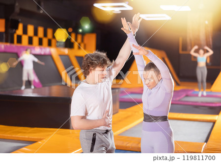 Man trainer instructing middle-aged woman doing exercises in trampoline arena Man trainer instructing middle-aged woman doing exercises in trampoline arena 119811700