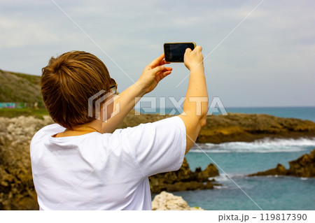 Middle age woman takes a photo on mobile phone on the Atlantic coast traveling through Spain, Santander. Female tourist with a short haircut in casual clothes on a seaside, seashore. Summer vacations. Middle age woman takes a photo on mobile phone on the Atlantic coast traveling through Spain, Santander. Female tourist with a short haircut in casual clothes on a seaside, seashore. Summer vacations. 119817390