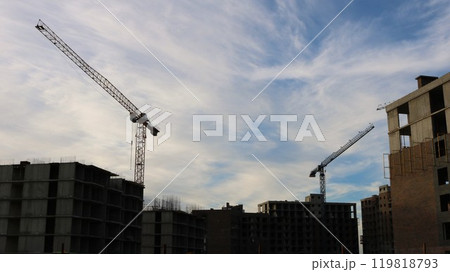 Behind unfinished multi-story buildings stand two construction cranes against blue cloudy sky, construction site in evening 119818793