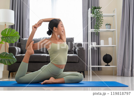 Sporty woman in green top and leggings stretching in Eka Pada Rajakapotasana exercise, One Legged King Pigeon pose on blue mat in bedroom at home.. Sporty woman in green top and leggings stretching in Eka Pada Rajakapotasana exercise, One Legged King Pigeon pose on blue mat in bedroom at home.. 119818797