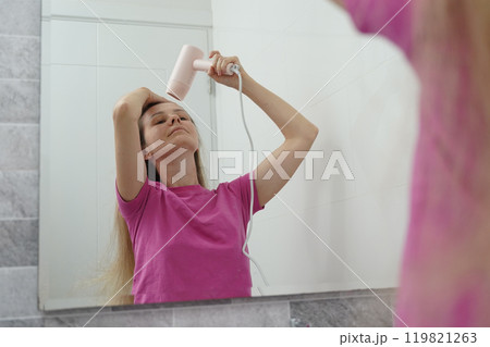 A woman is expertly styling her hair using a blow dryer and brush while in the bathroom A woman is expertly styling her hair using a blow dryer and brush while in the bathroom 119821263
