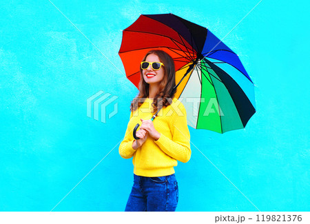 Happy smiling young woman holding colorful umbrella in autumn day on blue background Happy smiling young woman holding colorful umbrella in autumn day on blue background 119821376
