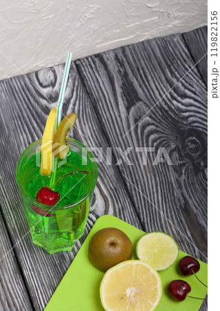 Green lemonade in a glass. Cherry floats in it. Garnished with a slice of lemon and kiwi. With a straw for a cocktail. Nearby on a cutting board are fruits for a cocktail. Green lemonade in a glass. Cherry floats in it. Garnished with a slice of lemon and kiwi. With a straw for a cocktail. Nearby on a cutting board are fruits for a cocktail. 119822156