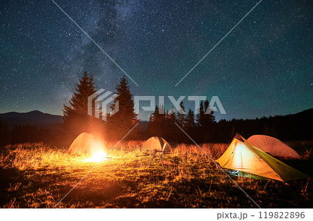 Several tents around glowing campfire on grassy clearing under starry sky with Milky Way. Warm light of campfire illuminates scene, creating cozy and serene night camping atmosphere in the mountains. Several tents around glowing campfire on grassy clearing under starry sky with Milky Way. Warm light of campfire illuminates scene, creating cozy and serene night camping atmosphere in the mountains. 119822896