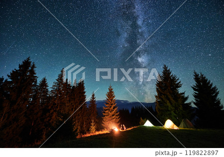 Night camping in mountains. Amidst forested mountain landscape, two tents glow warmly beside campfire under brilliant, star-filled sky. Silhouettes of tourists resting at campsite under Milky Way. Night camping in mountains. Amidst forested mountain landscape, two tents glow warmly beside campfire under brilliant, star-filled sky. Silhouettes of tourists resting at campsite under Milky Way. 119822897