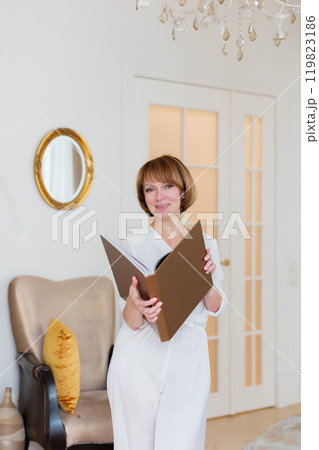 An adult beautiful woman spends time in her stylish living room and holds a big book in her hands. 119823186