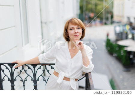 Adult woman in white suit is standing on balcony in town and looking at camera. 119823196