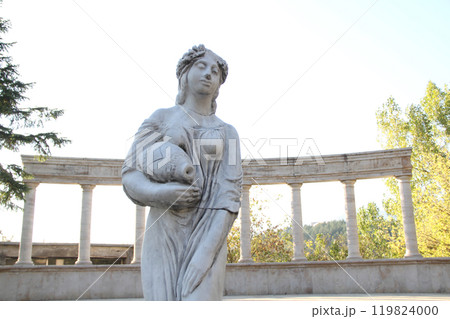 Marble statue of a woman holding an amphora in front of classical columns. 119824000