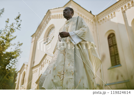 Low angle shot of African American priest in white robe holding wooden rosary walking in yard during warm day 119824614