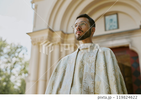 Low angle shot of young Caucasian priest dressed in white robe with golden patterns standing outside, beige church walls behind him 119824622