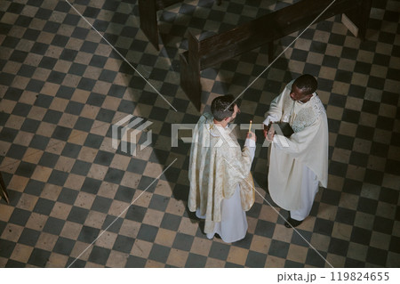 High angle shot of two unrecognizable priests in same robes holding candle and bible interacting in center of nave before church service 119824655