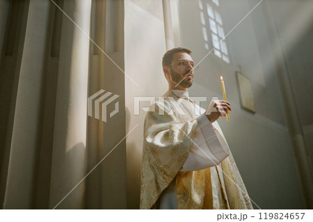 Low angle shot of ample church nave with young Caucasian priest in golden white robe holding long wax candle during service inside 119824657