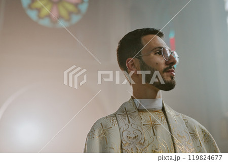 Low angle of young Caucasian priest in white robe with golden patterns standing in church full of light fog Low angle of young Caucasian priest in white robe with golden patterns standing in church full of light fog 119824677