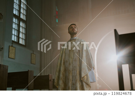 Low angle shot of young Caucasian priest dressed in white long robe with golden patterns looking at beautiful ceiling of church Low angle shot of young Caucasian priest dressed in white long robe with golden patterns looking at beautiful ceiling of church 119824678