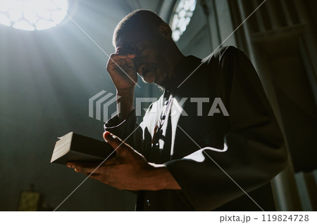 Low angle shot of African American priest in robe holding closed Bible crossing himself during church service 119824728