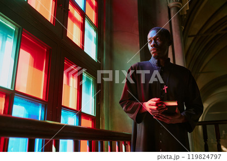 Low angle shot of African American priest with Bible looking out of multicolored stained window Low angle shot of African American priest with Bible looking out of multicolored stained window 119824797