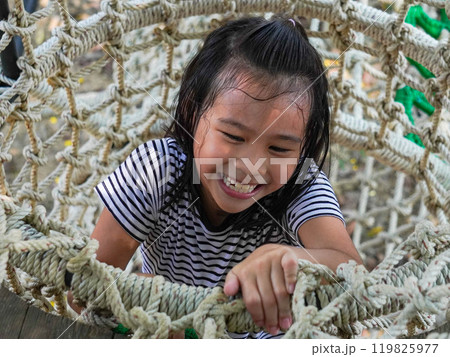 Children play outdoors on a wooden playground in a park. Happy girl playing on a rope net bridge in playground during summer. 119825977