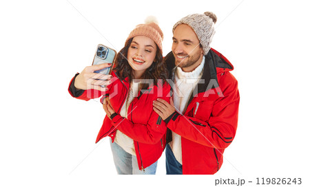 Young couple sitting on ice rink, smiling as they take selfie together, dressed in matching winter outfits. Love, togetherness, and winter joy. 119826243