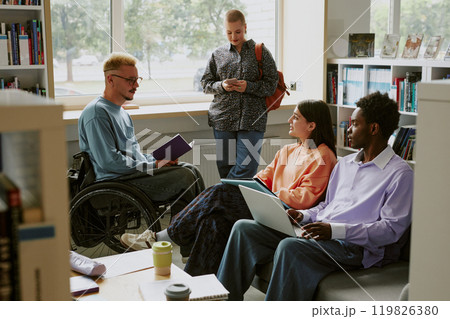 Group of diverse students discussing ideas in library with books and laptops surrounding them and one student in wheelchair holding notebook studying 119826380