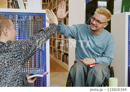 Two friends sharing high five in library surrounded by shelves with books while holding their own reads and smiling in wheelchair, showing camaraderie 119826384
