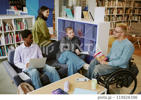 Group of diverse students studying in library, engaging in discussion with textbooks and laptops around. Bright, spacious study area surrounded by bookshelves 119826386