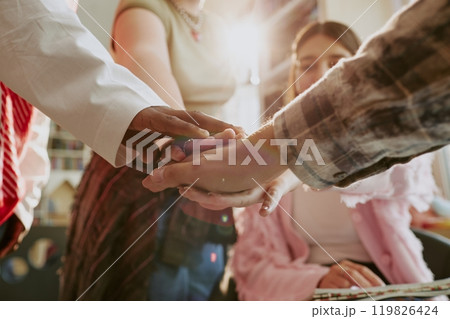 Hands of two individuals meeting in a friendly handshake with warm sunlight illuminating the scene in a lively room filled with background activity 119826424