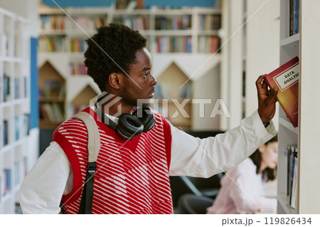 Young African American person selecting data analysis book in library, wearing red vest and headphones around neck. Bookshelf and other library users in background 119826434
