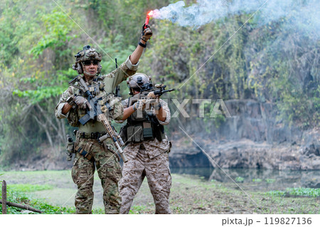 Two militaries or soldiers stay in battlefield near the river with one hold flare fireworks and one stand with point gun forward direction during outdoor practice. Two militaries or soldiers stay in battlefield near the river with one hold flare fireworks and one stand with point gun forward direction during outdoor practice. 119827136